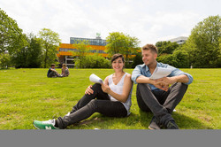 Zwei Studierende sitzen auf einer Wiese, im Hintergrund die Mensa