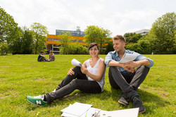 Zwei Studierende sitzen auf einer Wiese, im Hintergrund die Mensa