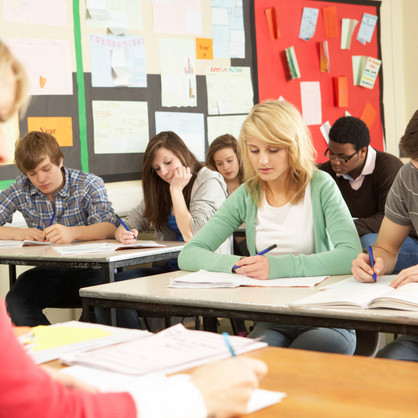 Schülerinnen und Schüler sitzen in einer Klasse und schreiben in Hefte, vor ihnen sitzt eine Lehrerin an einem Pult.