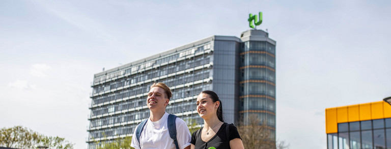 Ein junger Mann und eine junge Frau laufen über die Mensabrücke. Im Hintergrund sieht man den Mathetower mit TU-Logo und das Mensagebäude.