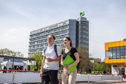 Ein junger Mann und eine junge Frau laufen über die Mensabrücke. Im Hintergrund sieht man den Mathetower mit TU-Logo und das Mensagebäude.