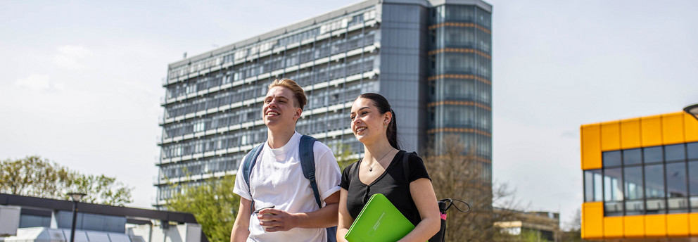 Studierende auf der Mensabrücke Ein junger Mann und eine junge Frau laufen über die Mensabrücke. Im Hintergrund sieht man den Mathetower mit TU-Logo und das Mensagebäude.