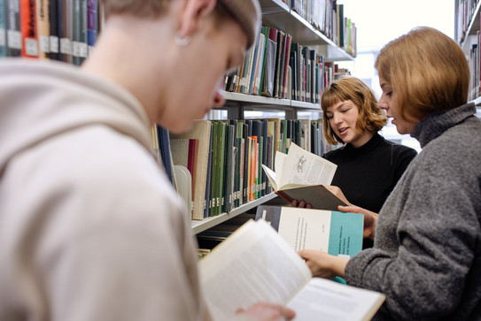 Drei Studierende stehen zwischen zwei Bücherregalen und lesen.