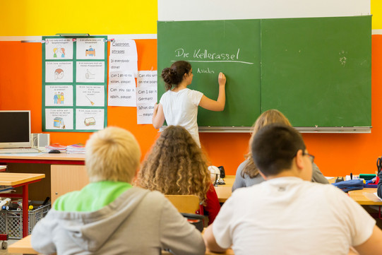 Klassenzimmer mit Tafel in einem Klassenzimmer sitzen Schüler*innen, eine junge Frau schreibt an der Tafel