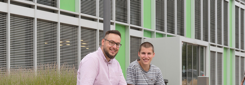Zwei Studierende sitzen vor einem Gebäude der Uni und lächeln in die Kamera. Vier weitere laufen im Hintergrund Richtung Gebäudeeingang.