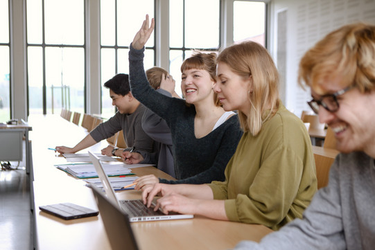 Studierende im Seminar Fünf Studierende sitzen in einem Seminarraum an Tischen. Eine Studentin hebt ihre Hand.