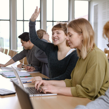 Fünf Studierende sitzen in einem Seminarraum an Tischen. Eine Studentin hebt ihre Hand.