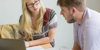 MatheHelpdesk Nahaufnahme von zwei Personen, die an einem Tisch sitzen. Die Person rechts hat Unterlagen vor sich liegen und spricht. Die Person links hört der anderen Person konzentriert zu.